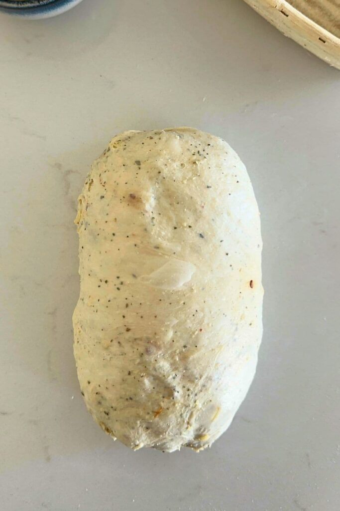 The dough of a sourdough Italian herb and cheese loaf that has been shaped into a batard and is ready to be lifted into a banneton.