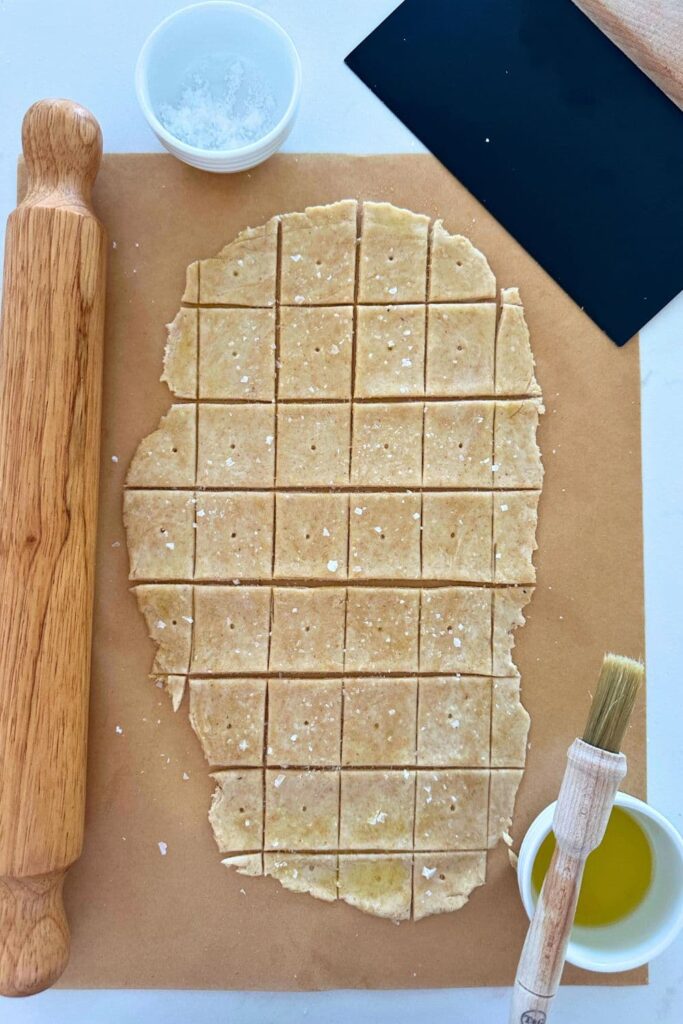 Whole wheat sourdough cracker dough that has been rolled out and cut into small squares. Each piece has a small hole in it. The dough has been brushed with olive oil and sprinkled with sea salt.