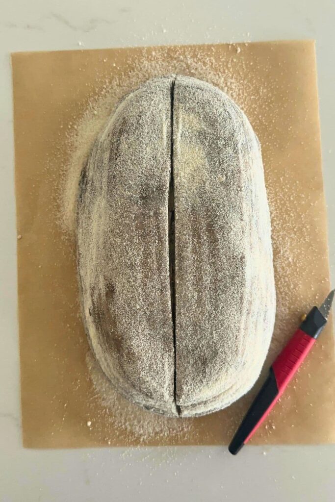 A loaf of sourdough cookies and cream bread tipped out of the banneton onto a piece of parchment paper and scored down the middle with a red handled lame.