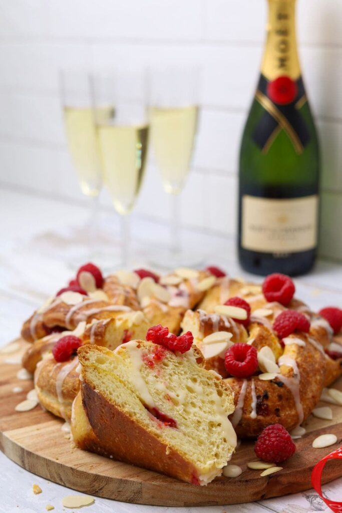 A sourdough brioche braided loaf decorated with flaked almonds and fresh raspberries displayed on a wooden board. There are 3 glasses of champagne and a bottle of Moet in the background of the photo.