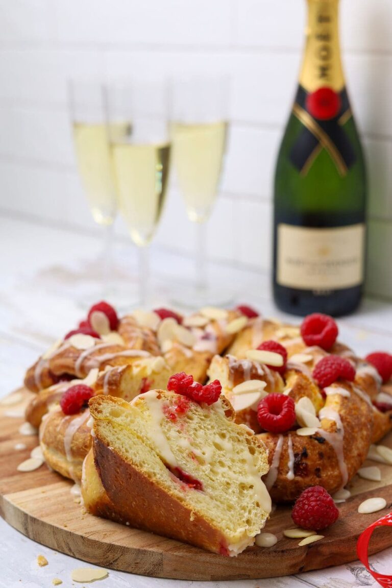 A sourdough brioche braided loaf decorated with flaked almonds and fresh raspberries displayed on a wooden board. There are 3 glasses of champagne and a bottle of Moet in the background of the photo.