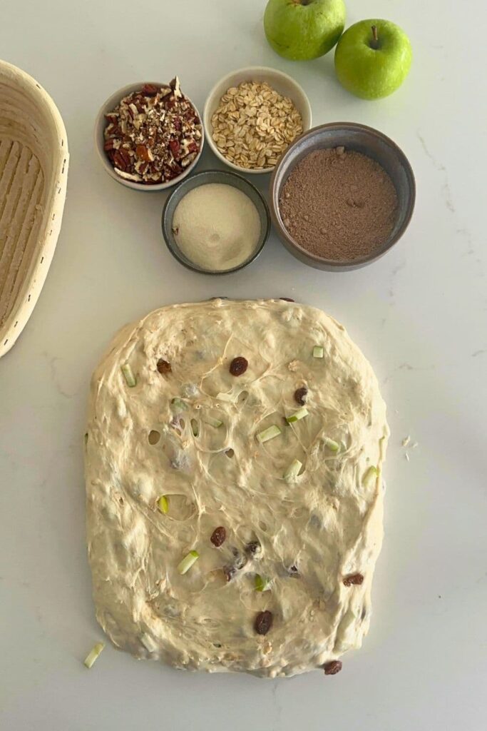 A rough rectangle of dough that is ready to shape into a batard. There are 4 bowls of inclusions ready to sprinkle over the dough. 2 green apples can also be seen in the photo but these are just there for decoration.