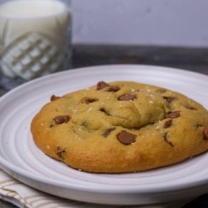 A huge single serve sourdough chocolate chip cookie sitting on a white plate. There is a glass of milk in the background.