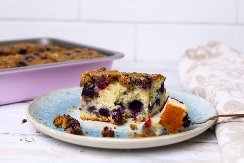 A horizontal image of a piece of sourdough blueberry buckle served on a blue stoneware plate. You can see a pink baking pan with the rest of the blueberry buckle in the background.
