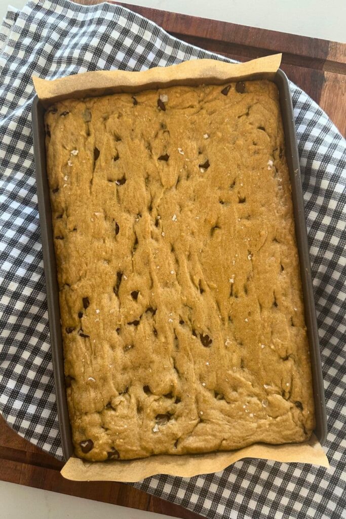 A tray of sourdough chocolate chip cookie bars sprinkled in sea salt that has been taken out of the oven and placed onto a black and white checked dish towel.