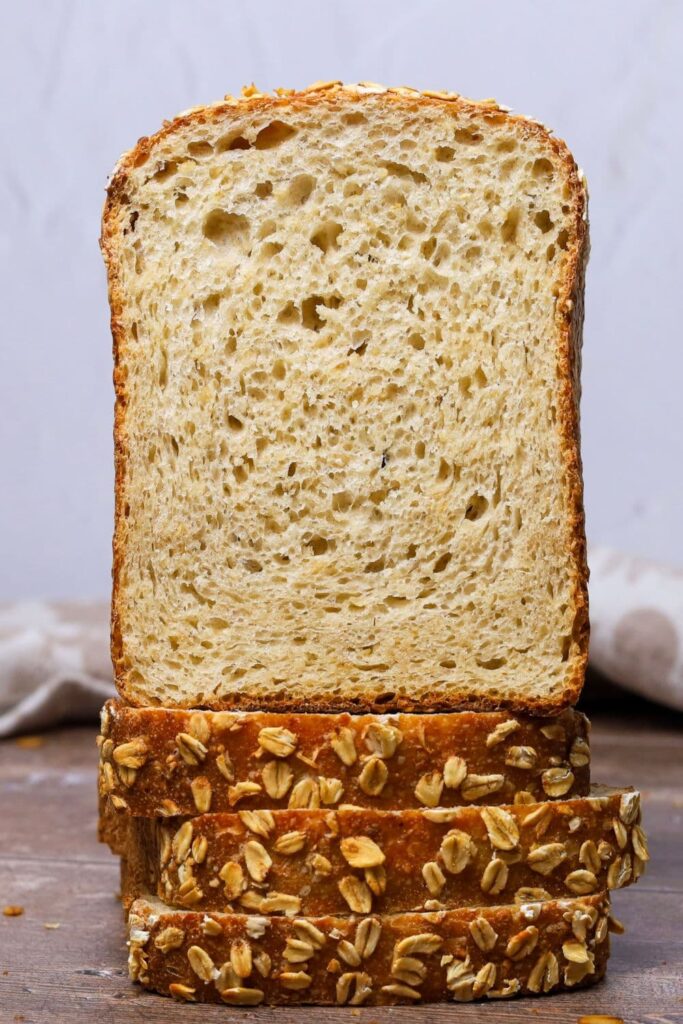 Half of a loaf of sourdough maple oat sandwich loaf sitting open faced on top of a stack of slices. You can see the soft crumb inside the loaf from the open face of the loaf. The slices are sitting so that you can see the oat crusted sides.