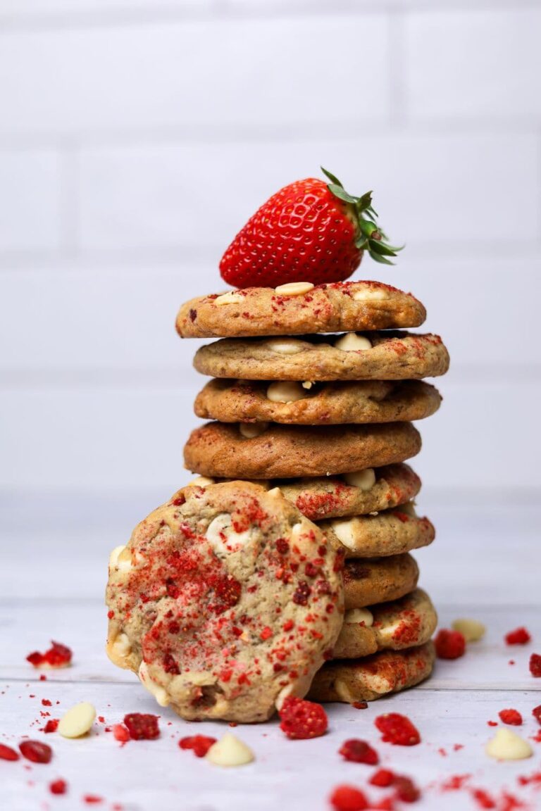 A stack of sourdough strawberries and cream cookies sitting on a white kitchen counter. One of the cookies is faced outwards so you can see the top of the cookie coated in freeze dried strawberries. There is a fresh strawberry sitting on top of the stack.