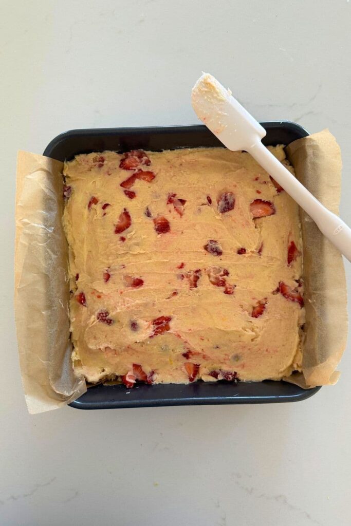 Sourdough strawberry bar batter added to a 9" x 9" baking pan. There is parchment paper overhanging the edges and a white spatula sitting on top.