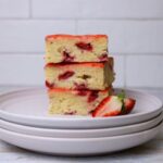 A stack of 3 sourdough strawberry bars sitting on a stack of 3 white plates. You can see the bright red strawberries and yellow lemon zest peaking through the golden crumb. There are some fresh strawberries garnishing the left hand side of the plate.