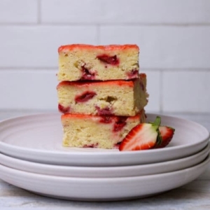 A stack of 3 sourdough strawberry bars sitting on a stack of 3 white plates. You can see the bright red strawberries and yellow lemon zest peaking through the golden crumb. There are some fresh strawberries garnishing the left hand side of the plate.