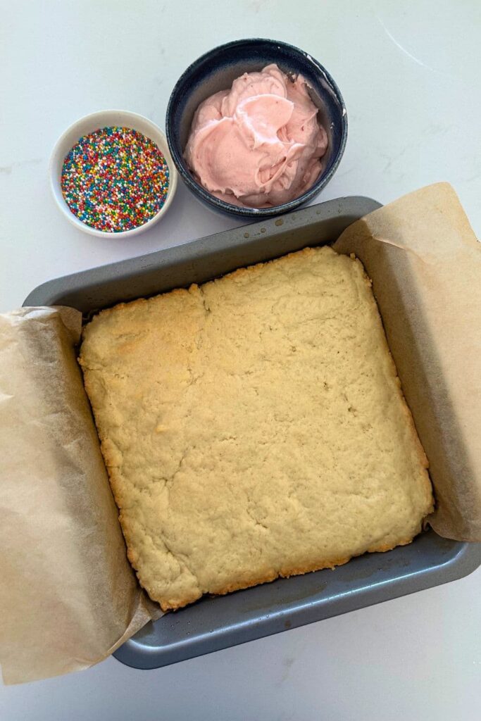 Sourdough sugar cookie bars that have been baked. They have been taken out of the oven to cool in the pan. There is a bowl of strawberry frosting and rainbow frosting in the photo as well.