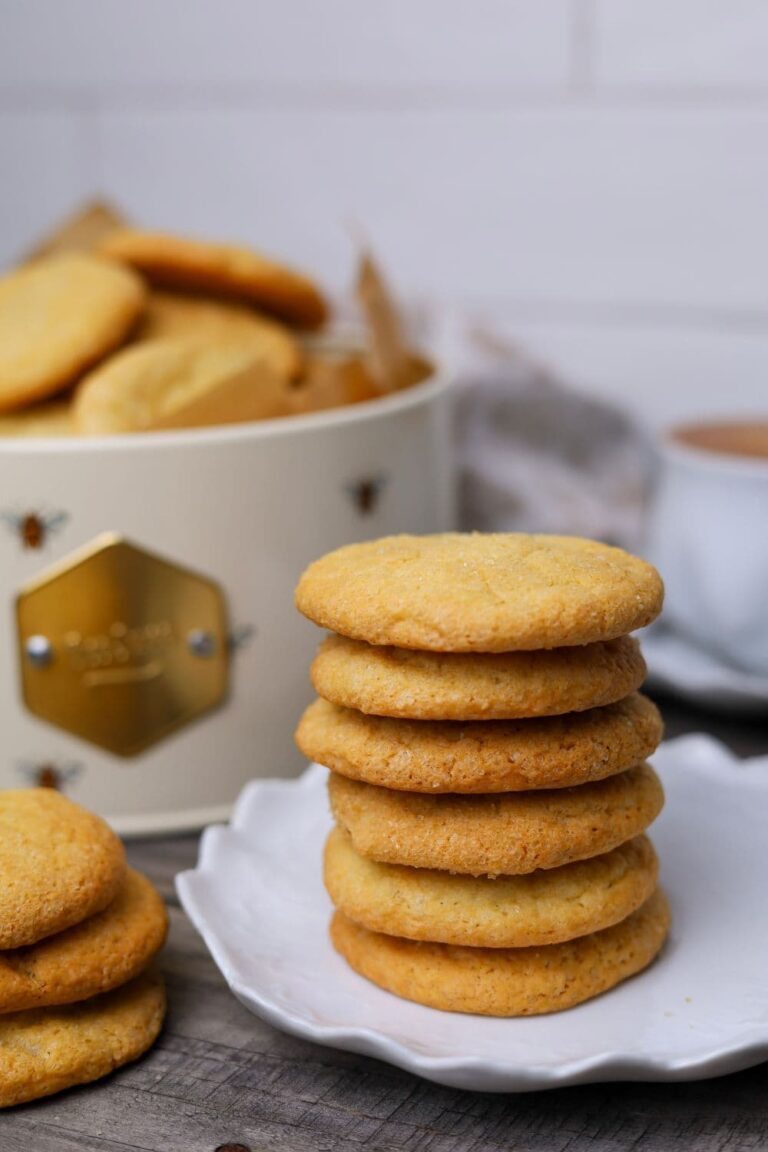 A stack of 6 sourdough vanilla wafer cookies sitting on a white saucer. There is a smaller stack of three cookies to the left of this stack and a large cream colored tin of cookies and a cup of coffee in the background.