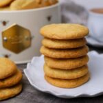 A stack of 6 sourdough vanilla wafer cookies sitting on a white saucer. There is a smaller stack of three cookies to the left of this stack and a large cream colored tin of cookies and a cup of coffee in the background.
