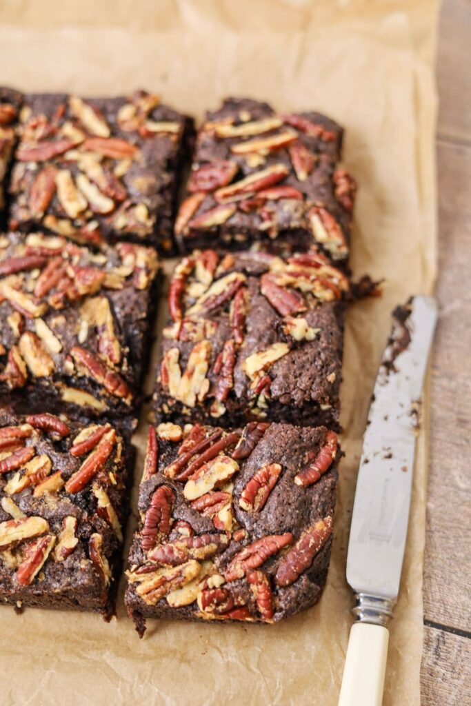 6 squares of spiced sourdough brownies topped with buttered pecans sitting on a sheet of parchment paper. There is a butter knife sitting next to the brownies.
