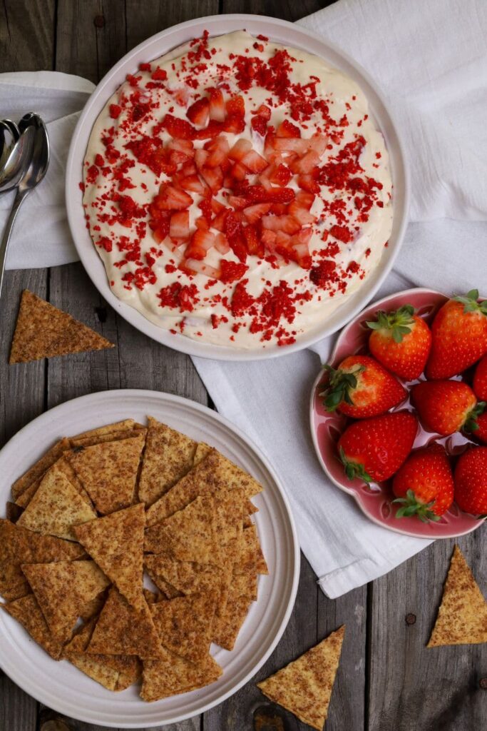 A white dish of strawberry cheesecake dip garnished with fresh and freeze dried strawberries sits at the top of the photo. There is another white dish piled with sourdough cinnamon sugar crackers lower down and then a small pink dish of strawberries to the right of the photo.