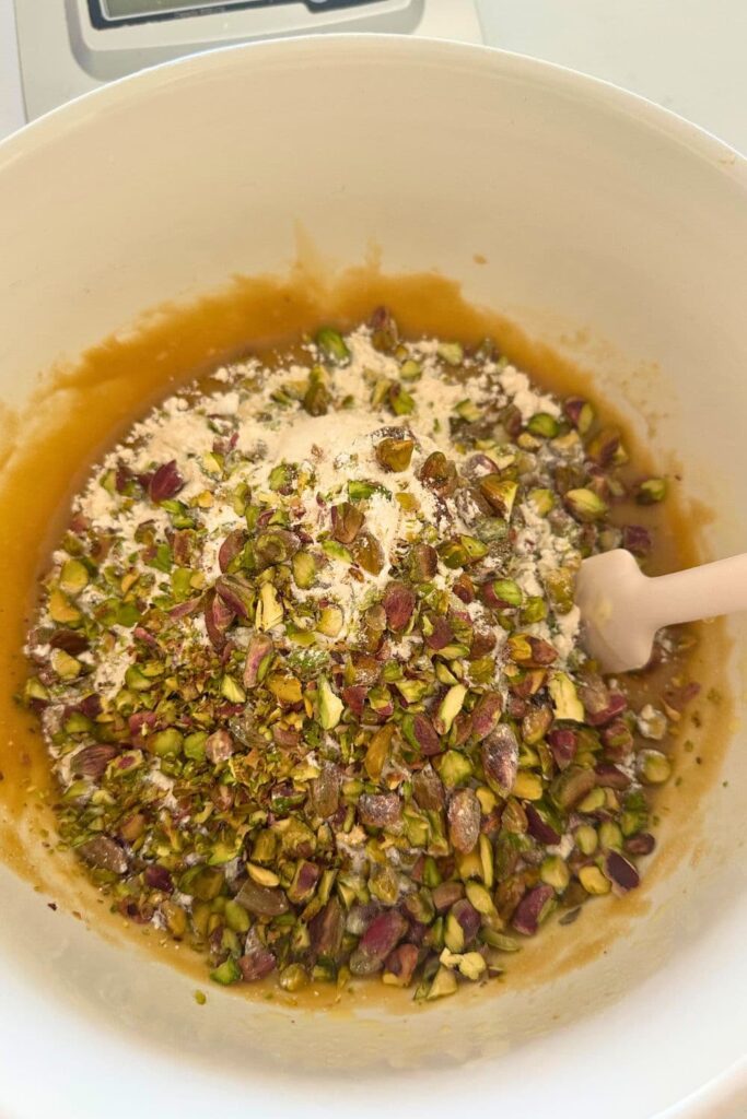Dry ingredients sitting on top of wet ingredients. You can see the purple and green colors of the pistachios. There is a white spatula in the bowl ready to stir the wet and dry ingredients together.