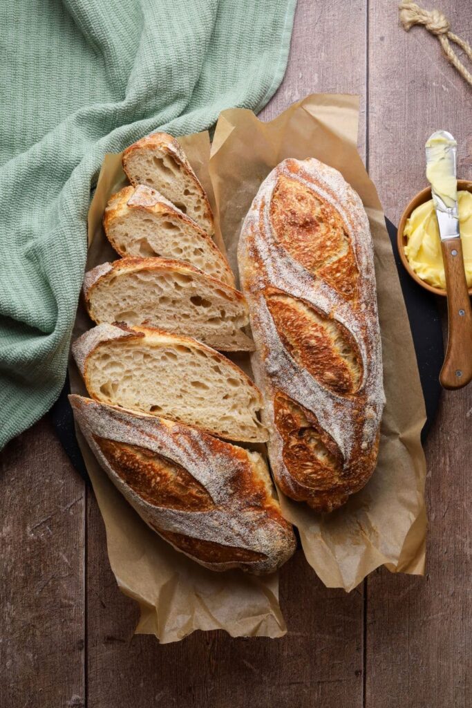 A sourdough batard that has been sliced into two halves and then baked. The two baked loaves are sitting on some parchment paper. The left hand loaf has been sliced up, the right hand loaf is whole.