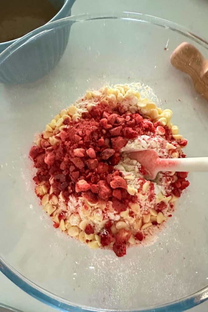 A bowl of dry ingredients necessary to make sourdough strawberries and cream cookies. You can see the bright red freeze dried strawberries and creamy white chocolate in the bowl.