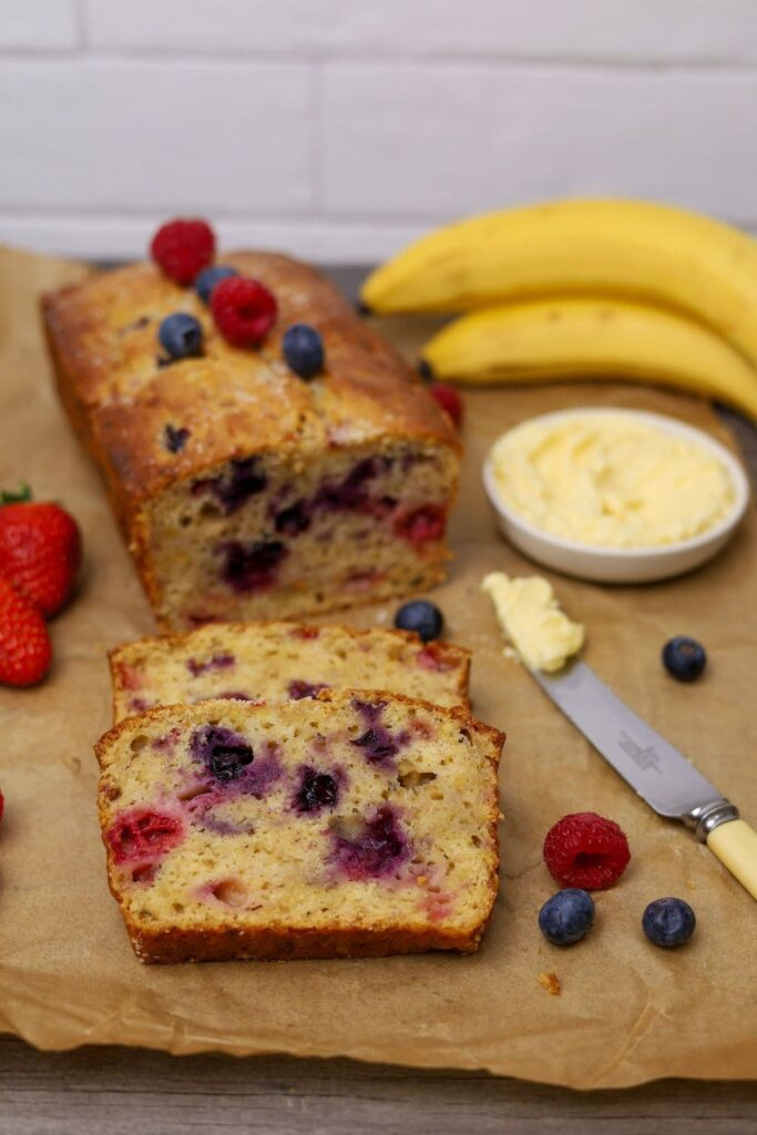 A loaf of sourdough berry banana bread that has been sliced and laid out on a sheet of parchment paper so you can see the colorful berries inside. There is a small dish of butter and a butter knife, as well as some yellow bananas in the photo.