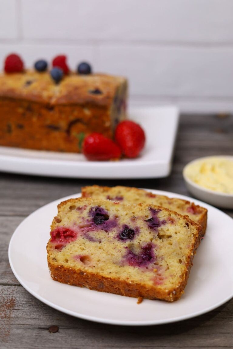 A feature image of sourdough berry banana bread. There are two slices of sourdough berry banana bread displayed on a white plate. You can see the rest of the loaf in the background of the photo garnished with fresh berries.
