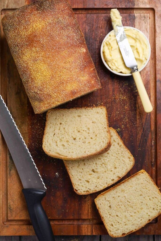 Sourdough English muffin loaf on wooden cutting board with slices and a bowl of butter.