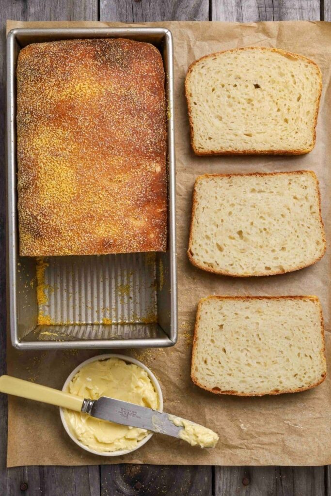 Sourdough English muffin loaf on parchment paper with three slices of bread next to a bowl of butter with a butter knife.