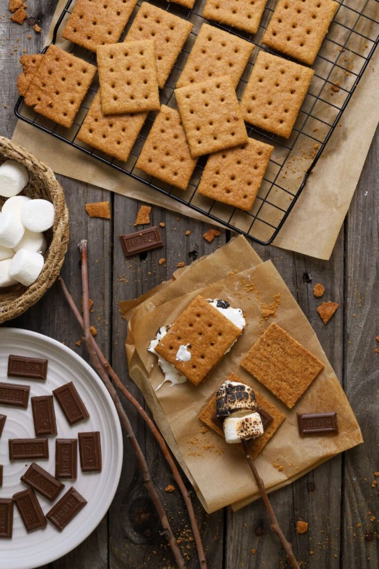 A photo taken from above showing a black wire rack of sourdough graham crackers at the top of the photo. There is also a small basket of white marshmallows and a white plate of Hershey's pieces. To the right of the photo there is a piece of parchment paper where some s'mores have been made with toasted marshmallows and sticks.
