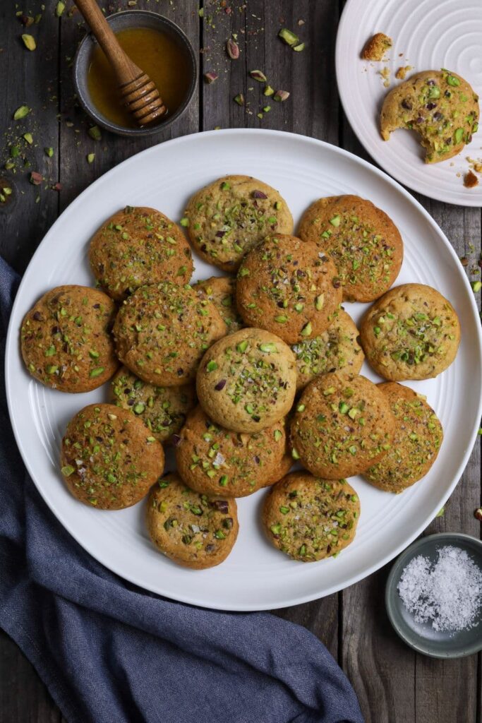 A large round white plate with a selection of sourdough honey pistachio cookies laid out on it. There is a small dish of honey at the top of the photo, as well as a small bowl of flakey sea salt at the bottom next to a denim blue dish towel.