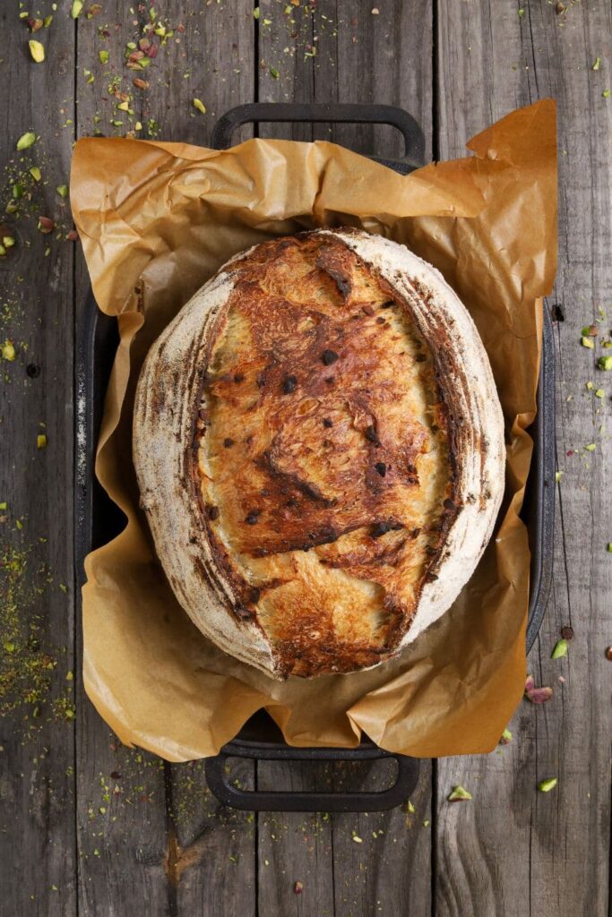 A whole loaf of sourdough pistachio bread that is sitting in a cast iron bread pan. The bread has been baked and is photographed from above to show the whole loaf.