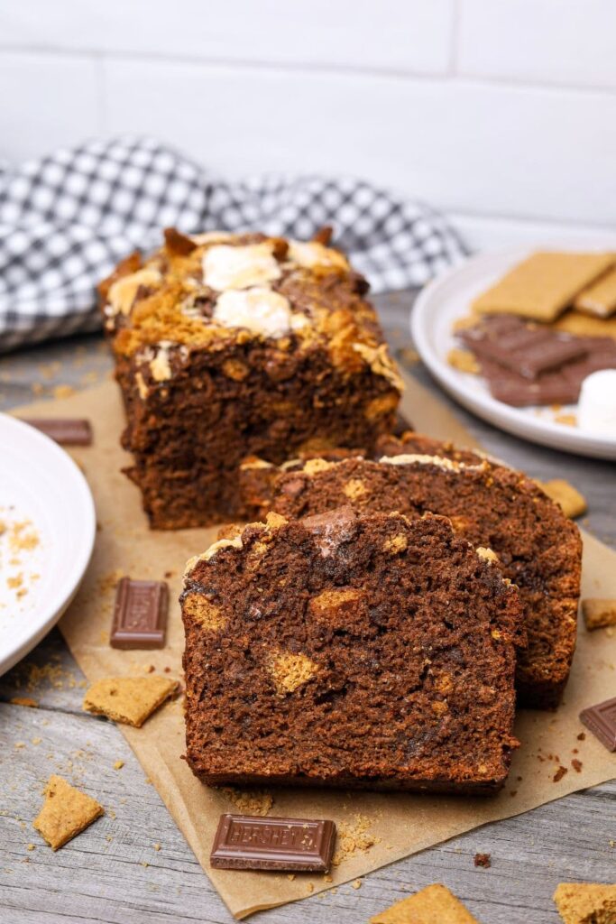 A close up photo of 2 slices of sourdough s'mores banana bread surrounded by Hershey's pieces and sourdough graham crackers. There is also a black and white dish towel in the background of the photo.