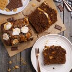 A flat lay photo of sourdough s'mores banana bread taken from above. There is a loaf of sourdough s'mores banana bread that has been sliced up so you can see the crumb inside. A piece of the loaf is sitting on a white plate.
