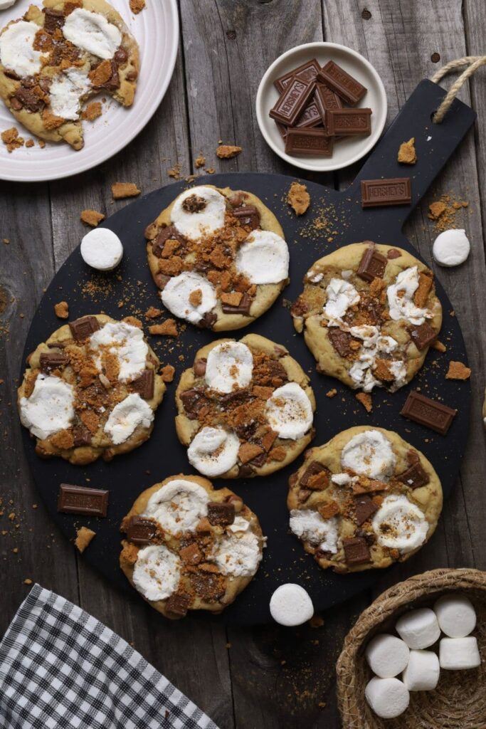 6 sourdough s'mores cookies sitting on a round black serving board. There are some decorative items surrounding the board, but these are ingredients that have been used in the sourdough s'mores cookies.