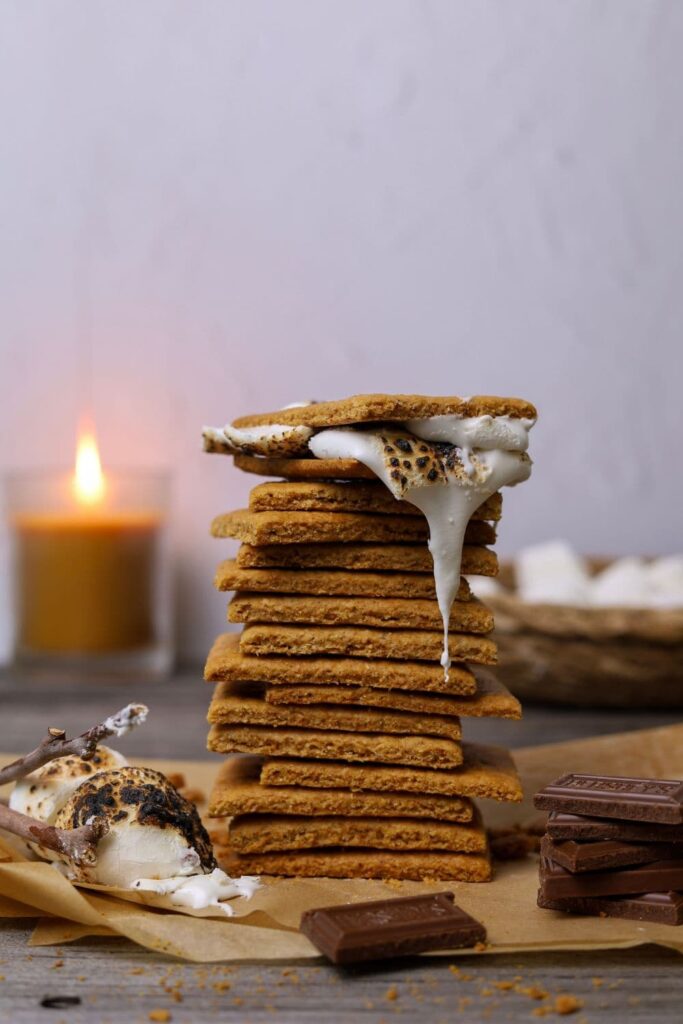 A stack of 15 sourdough graham crackers with a melted marshmallow dripping down from the top of the stack. There is a candle in the background. You can see some toasted marshmallows and Hershey's chocolate pieces in the foreground of the photo.
