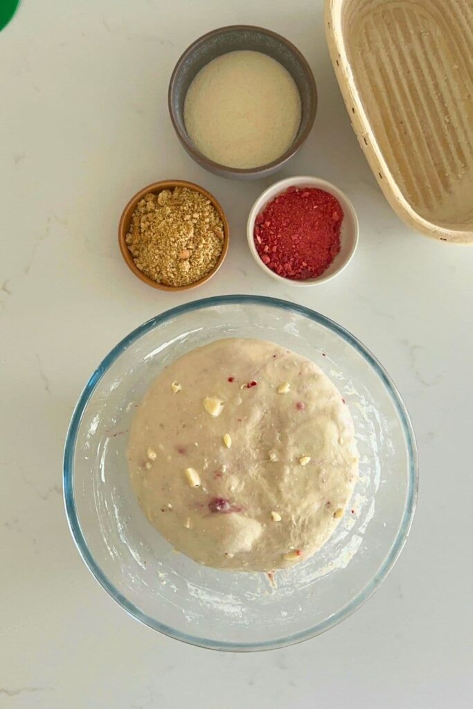 A glass bowl of dough that has been bulk fermented until doubled. There are 3 small bowls above the larger bowl containing semolina, cookie crumbs and freeze dried strawberries.
