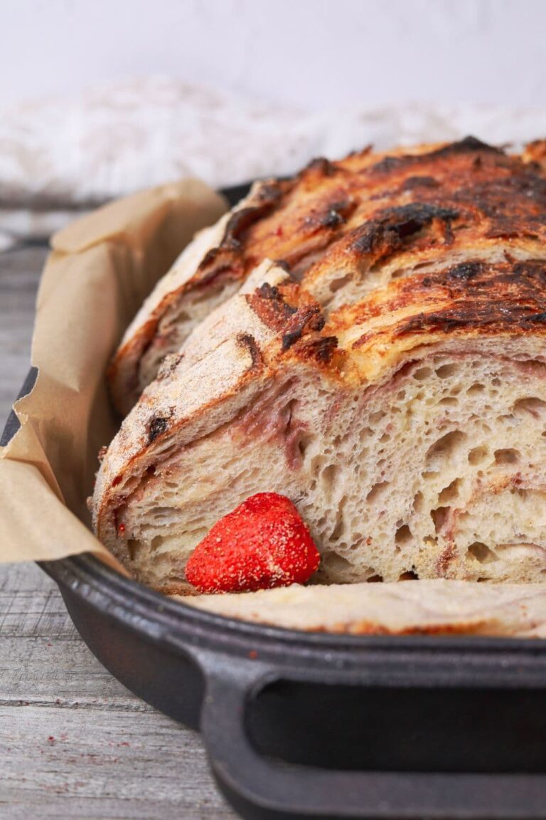 A close up photo of a sourdough strawberries and cream loaf. You can see the freeze dried strawberries and creamy white chocolate swirled through the open crumb. There is a freeze dried strawberry garnishing the loaf.