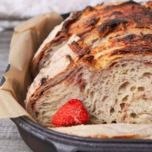A close up image of a loaf of strawberries and cream sourdough bread. You can see the pink streaks of freeze dried strawberries stained through the crumb. There is a whole freeze dried strawberry garnishing the bread. It is sitting in a black cast iron bread pan.