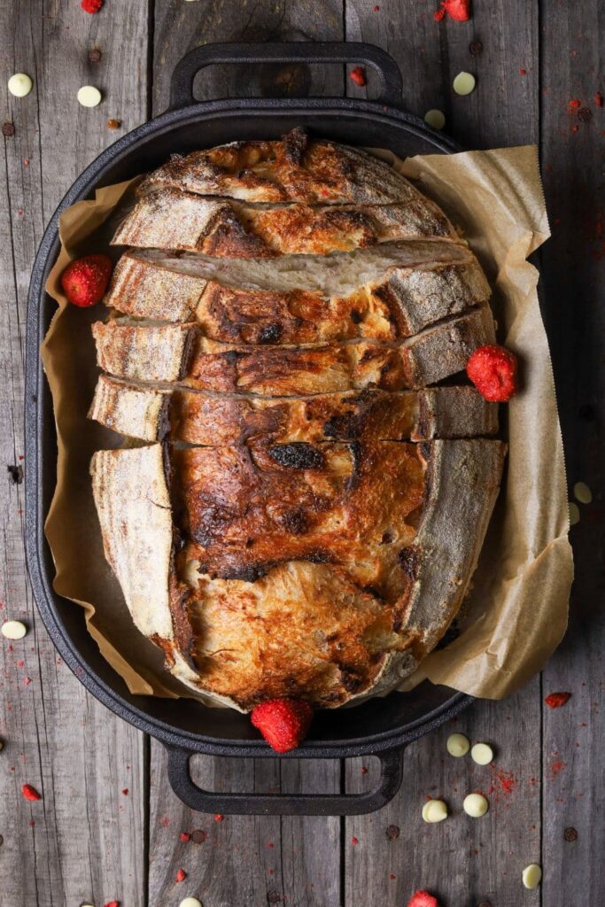 A loaf of sourdough strawberries and cream bread that has just been baked and is displayed in a black cast iron bread pan. There are 3 freeze dried strawberries garnishing the loaf.