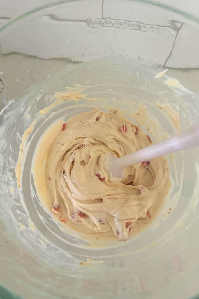A glass Kitchenaid bowl containing the strawberry cheesecake dip that has just had the diced fresh strawberries folded through with a white spatula.