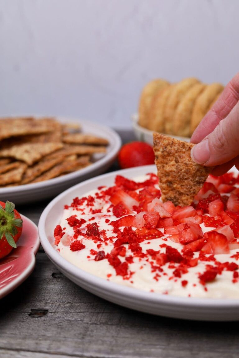 Strawberry cheesecake dip garnished with fresh and freeze dried red strawberries served on a white plate. There is a hand dipping a sourdough cracker into the dip.
