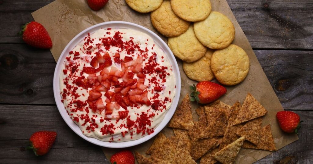 A white plate containing strawberry cheesecake dip garnished with red strawberries. The white plate sits on a wooden board and is surrounded with wafer biscuits and crackers and fresh strawberries.
