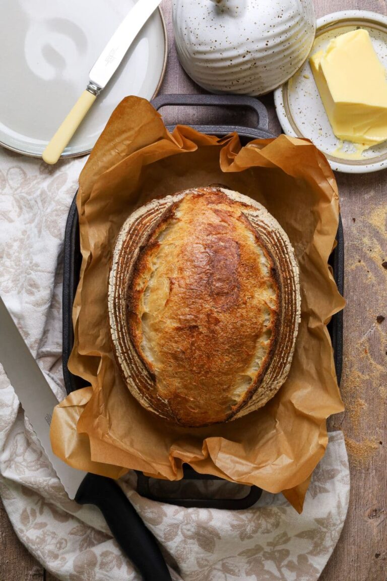 A loaf of crusty sourdough bread nestled in a piece of parchment paper inside a cast iron bread pan. There is a black bread knife, a side plate and a butter dish surrounding the loaf.