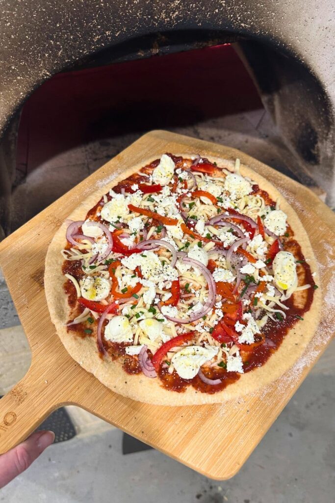 A whole wheat sourdough pizza base topped with cheese and vegetables being put into a wood fired pizza oven using a wooden pizza peel.