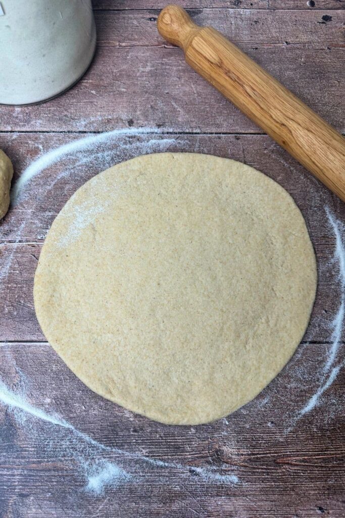 A whole wheat sourdough pizza crust rolled out on a wooden surface. There is a rolling pin at the top of the photo.