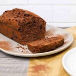 double chocolate sourdough banana bread loaf on a plate with one slice cut, dusted with cocoa powder and served on parchment paper.