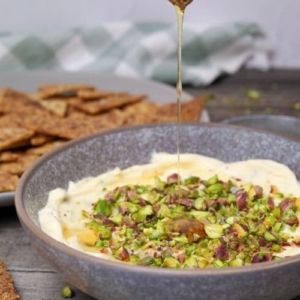 Zoom in of honey pistachio cream cheese in serving bowl with honey being poured on top and a serving tray with cinnamon sugar crackers in background.