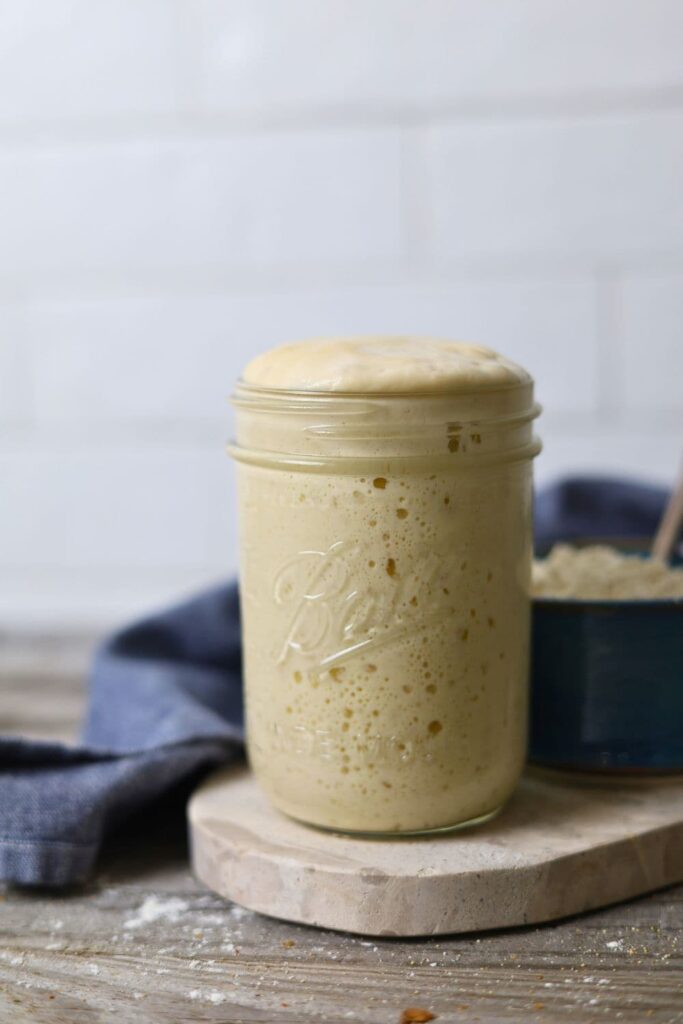 A mason jar filled with sourdough starter that has risen all the way to the top of the jar and is about to over flow. The jar is sitting on a marble board. There is a blue dish towel behind the jar.