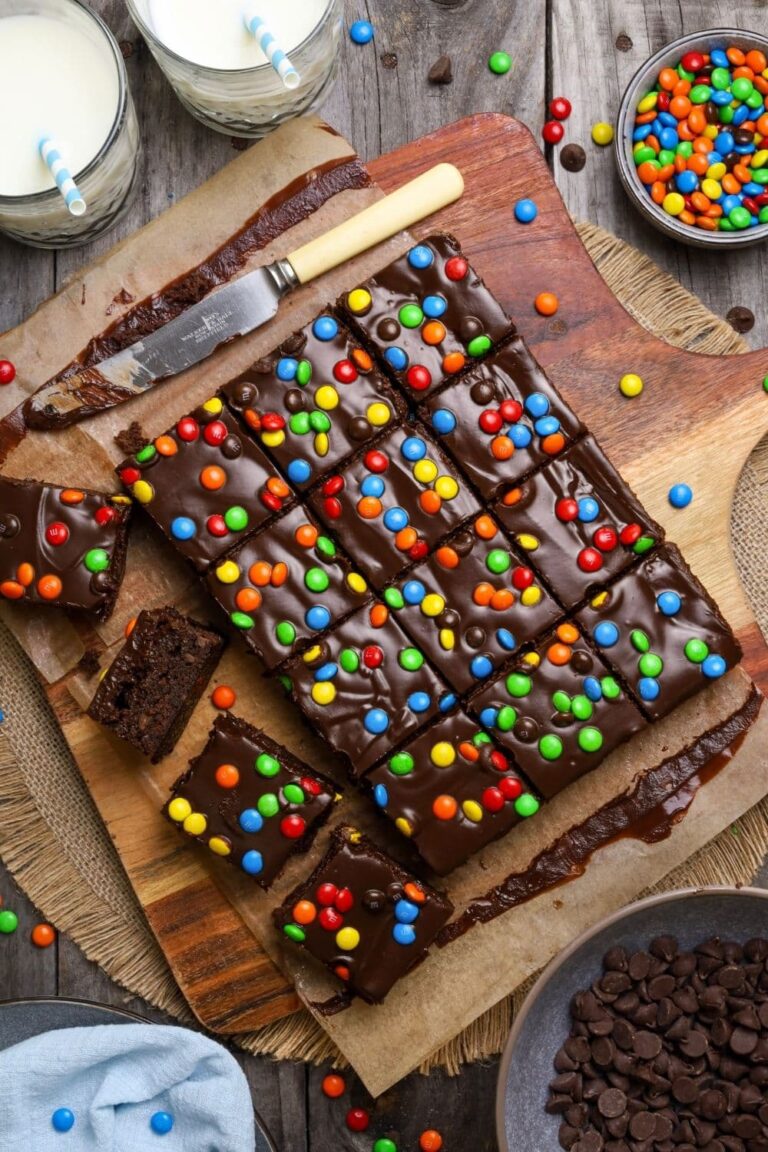 Sourdough cosmic brownies sliced on a cutting board over a placemat, next to a bowl of chocolate chips, candies and two glasses of milk.