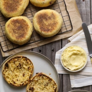 sourdough discard cinnamon raisin English muffins on cooling rack next to a plate with an English muffin sliced open.
