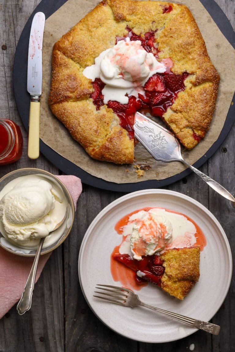 A sourdough strawberry galette topped with vanilla ice cream. There is a pretty cake slice sitting alongside the galette and a slice has been placed on a white plate and drizzled with strawberry sauce.