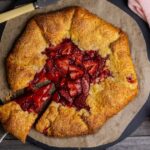 A vertical photo of a sourdough discard strawberry galette brimming with red strawberries. A slice has been taken out of the galette, but is still sitting on the plate. There is a bowl of vanilla ice cream sitting on a pink linen napkin above the galette and a small jar of strawberry sauce.
